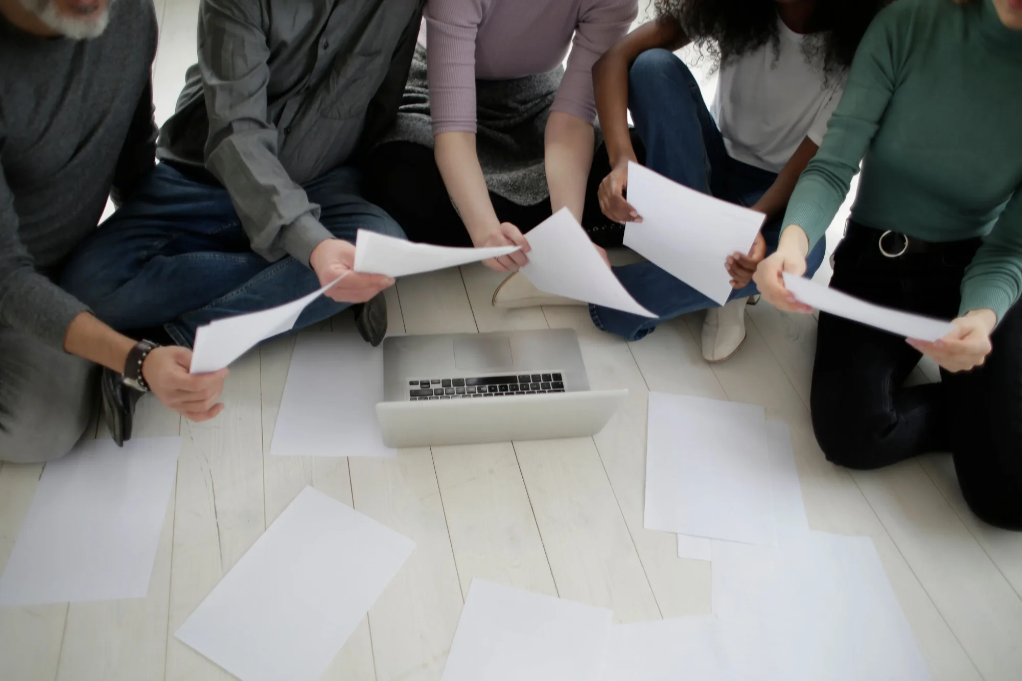 Diverse team sitting on floor reviewing document pages around a laptop for knowledge sharing